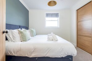 A neatly made bed with white linens and a few pillows, a window with a patterned valance, and a wooden door. - Firthview | Crail (Anstruther)