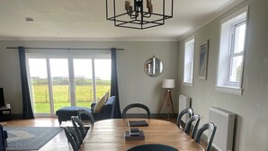 A dining room with a wooden table and chairs, a chandelier, and a view through large windows. - Firthview | Crail (Anstruther)