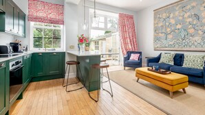 A modern kitchen with green cabinets, a central island with stools, a dining area with a blue sofa and yellow ottoman, and a large floral mural on the wall. - Greyfriars Bothy (St Andrews)