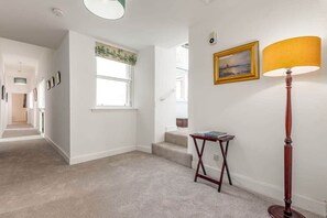 A hallway with carpeted flooring, a staircase, a small table with a lamp, and framed pictures on the wall. - The Cross Keys Apartment (85D Market Street) (St Andrews)