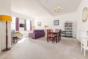 A spacious living room with a purple sofa, a dining table with chairs, a bookshelf, and a large mirror on the wall. - The Cross Keys Apartment (85D Market Street) (St Andrews)