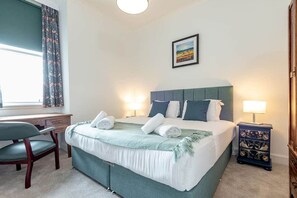 A neatly made bed with pillows and a blanket, a desk with a chair, a bedside table with a lamp, and a window with curtains. - The Cross Keys Apartment (85D Market Street) (St Andrews)