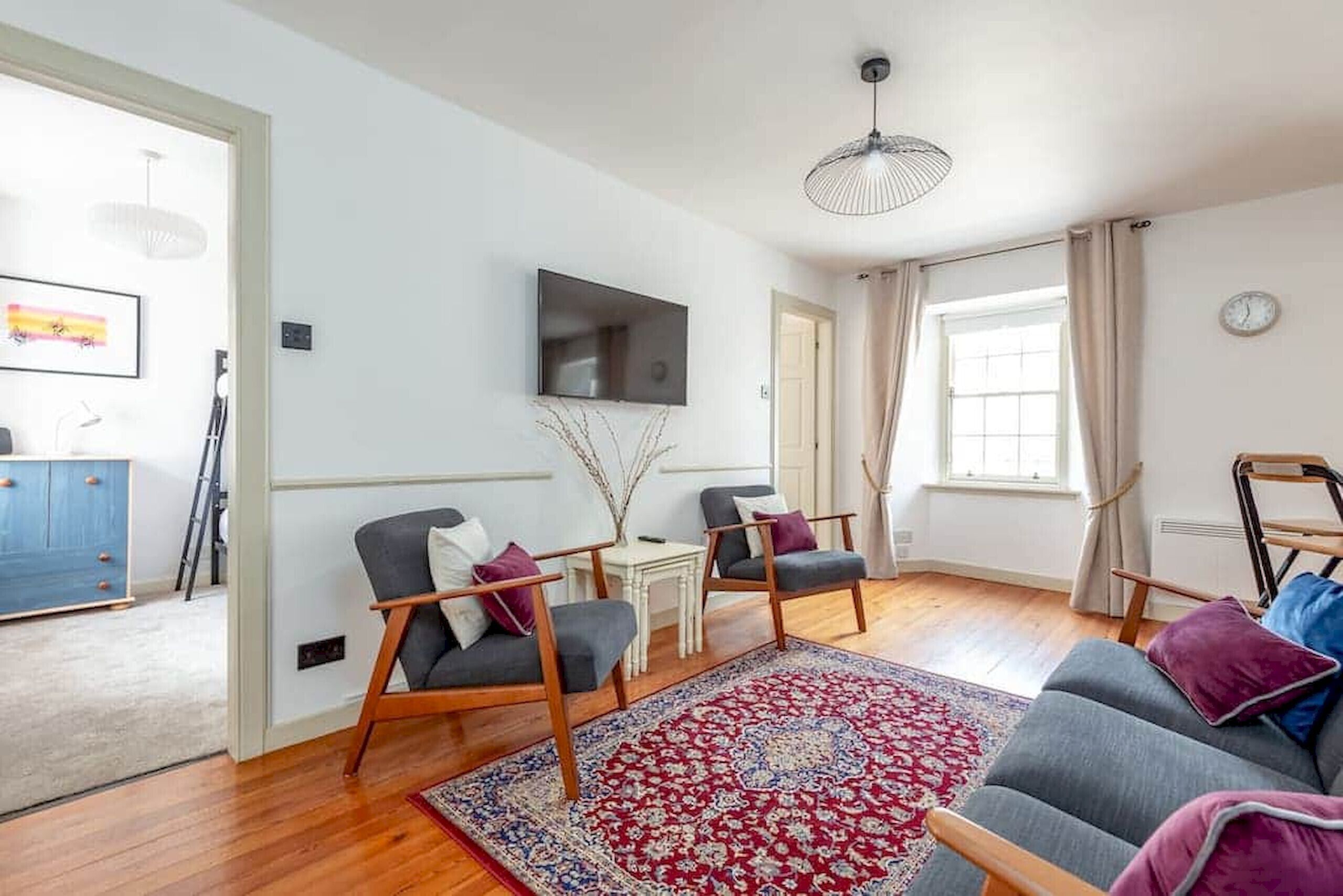 A modern living room with a grey sofa, two armchairs, a red patterned rug, a TV mounted on the wall, and a wooden floor.