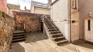 A narrow alleyway with a stone wall on one side and a building with a white door on the other. There is a set of concrete steps leading up to a metal staircase. - Castle Loft (St Andrews)