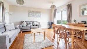 A modern living room with a grey sofa, a wooden dining table with chairs, a white rug, and a large window with curtains. - Bell Rock Cottage (St Andrews)
