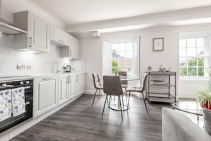 A modern kitchen with white cabinetry, a dining area with a glass table and chairs, and a view of the city through large windows.