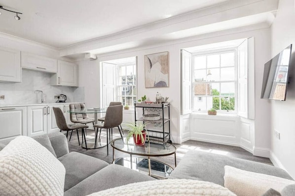 A modern living room with a grey sofa, a glass coffee table, and a dining area with chairs and a small table.