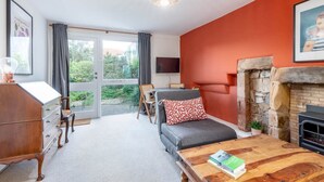 A cozy living room with a fireplace, a sofa, a coffee table with books, a TV, and a large window with curtains. - The Cooperage Garden Apartment | Crail (Crail)