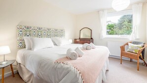 A bedroom with a large bed, a bedside table, a chair, a window with curtains, and a wall with a floral pattern. - The Cooperage Garden Apartment | Crail (Crail)