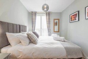 A neatly made bed with a grey headboard, white bedding, and a grey throw blanket. There are two white pillows, a grey pillow, and a pair of white towels on the bed. A modern pendant light hangs above the bed. - The Argyle Apartment (St Andrews)