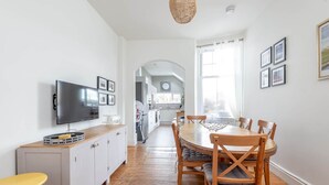 Une cuisine moderne avec des meubles blancs, une table à manger en bois avec des chaises, un téléviseur fixé au mur et une fenêtre avec des rideaux.