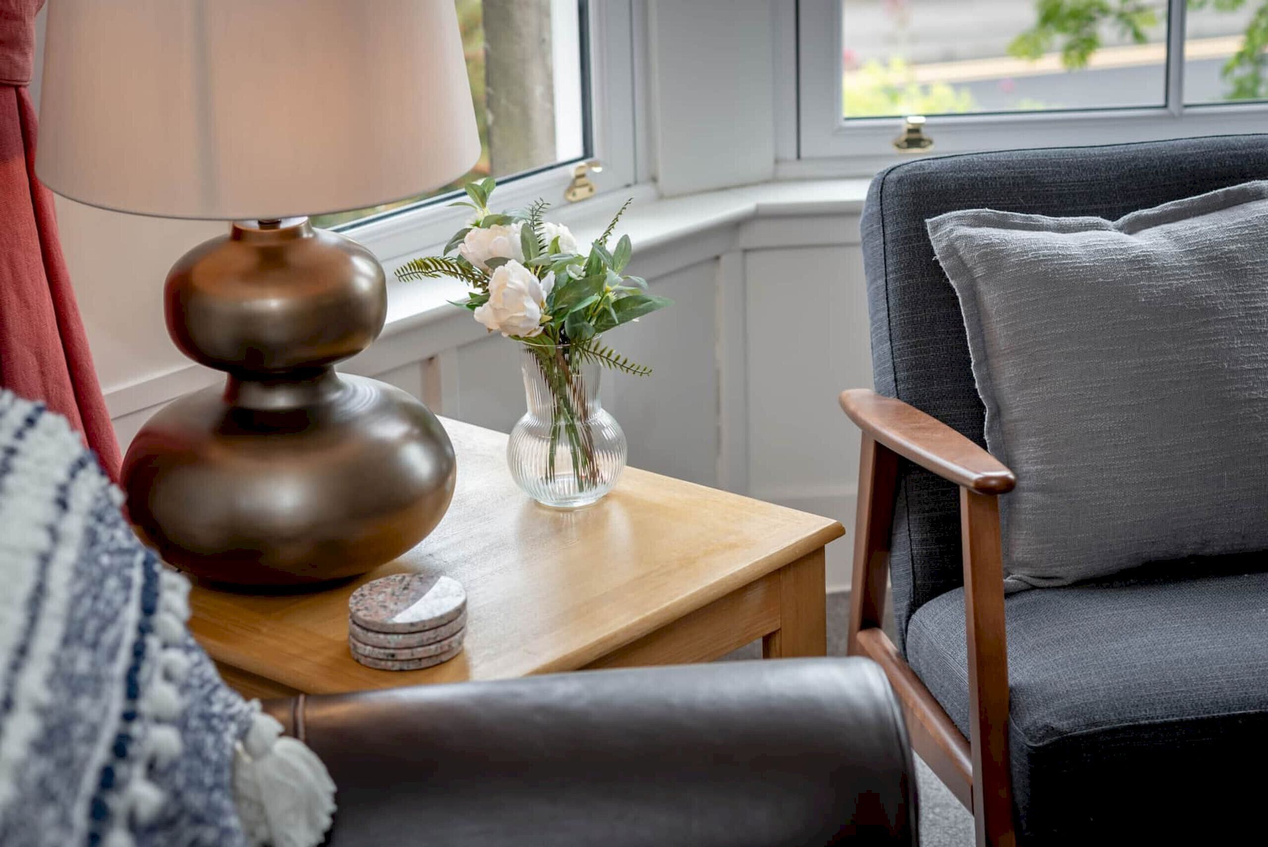 A cozy room with a wooden side table, a lamp, a vase with flowers, and a grey upholstered chair.