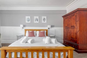 Cama bem arrumada com lençóis brancos e almofadas cor-de-rosa, cabeceira de madeira e um banco de madeira aos pés da cama. Três peças de arte emolduradas acima da cama, e um guarda-roupa de madeira à direita.