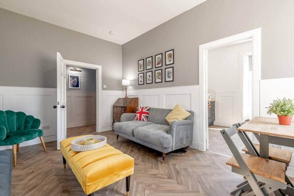 Uma sala de estar com um sofá cinzento, um banquinho amarelo, uma poltrona verde, uma mesa de jantar de madeira com uma planta em vaso, e uma porta que dá para outro cômodo.