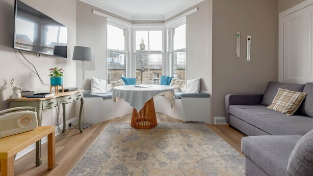 A cozy living room with a bay window, a flat-screen TV, a dining table, and a grey sofa. - Argyle Street Apartment (No 44) (St Andrews)