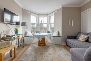 A cozy living room with a bay window, a flat-screen TV, a dining table, and a grey sofa.