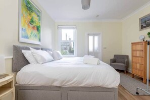 Un dormitorio con una cama grande, un cabecero gris, un sillón gris, un armario de madera y un cuadro en la pared.