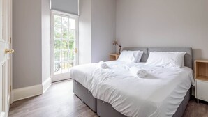 A bedroom with a large bed, a nightstand, a lamp, and a window with a view of greenery. - Queen's Gardens (No 18B) (St Andrews)