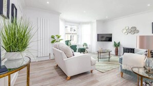 Een moderne woonkamer met een witte bank, een bijzettafeltje en een aan de muur bevestigde televisie. Er staan planten en er staat een groene fauteuil in de kamer.