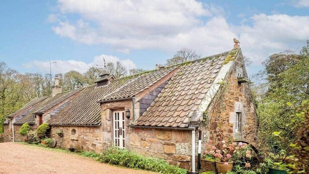 A stone cottage with a tiled roof, surrounded by greenery and a gravel path. - Park Mill Cottages | The Granary (St Andrews)