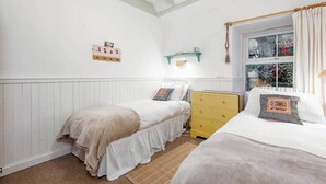 A bedroom with two beds, a yellow dresser, a window with curtains, and a wall-mounted shelf. - Park Mill Cottages | Mill Cottage (St Andrews)