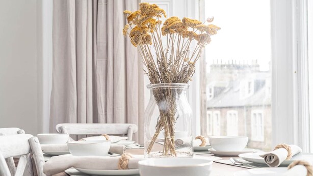 A dining table set with white plates, bowls, and cutlery, a vase with dried flowers, and a view of buildings through a window. - Skye Sands | Alexandra Penthouse (No 11) (St Andrews)