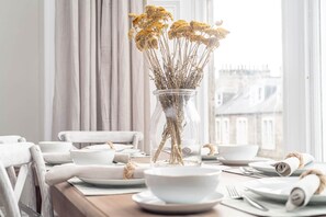A dining table set with white plates, bowls, and cutlery, a vase with dried flowers, and a view of buildings through a window. - Skye Sands | Alexandra Penthouse (No 11) (St Andrews)