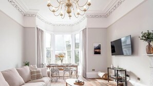 A spacious living room with a large sofa, a dining table, and a chandelier. - Skye Sands | Alexandra Penthouse (No 11) (St Andrews)