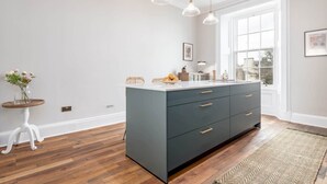 A kitchen with a large island, white walls, and wooden flooring. There are pendant lights above the island and a window providing natural light. - Skye Sands | Alexandra Penthouse (No 11) (St Andrews)