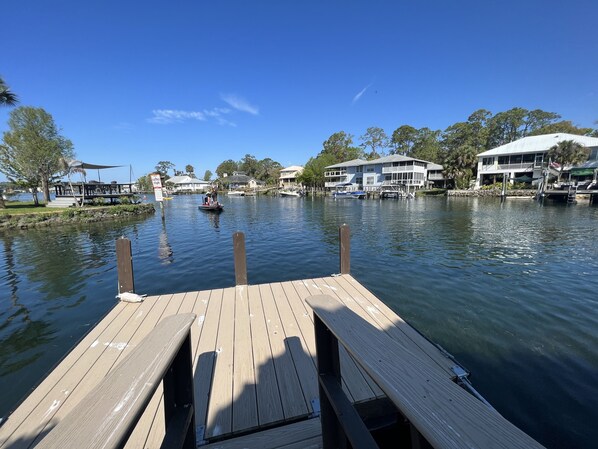 A wooden dock extending into calm water with a boat in the distance, surrounded by houses and trees.