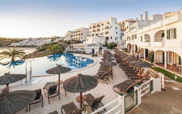 A resort pool area with lounge chairs and umbrellas, surrounded by white buildings with balconies. - Beachfront Apartment with Stunning Sea Views (Arenal d'en Castell)