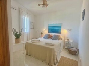 A bedroom with a bed, bedside tables, a hanging plant, a painting, and a ceiling fan.