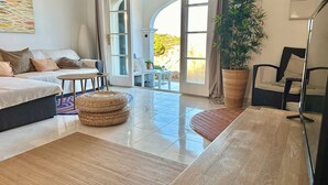 En moderne stue med sofa, sofabord og spisebord. Der er et stort vindue med udsigt til grønne omgivelser.