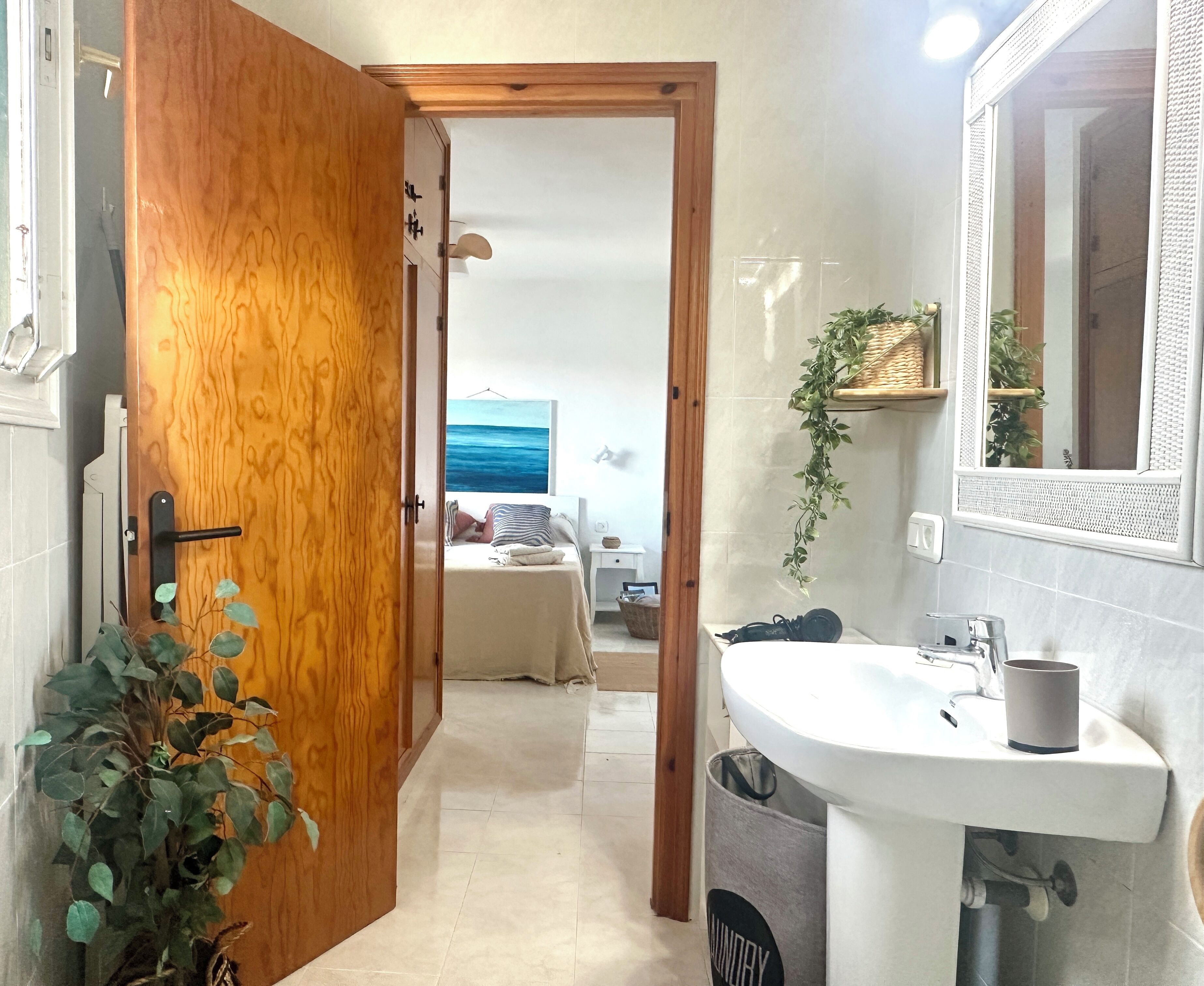 Een smalle gang leidt naar een slaapkamer met een bed, een nachtkastje en een wandplank met planten. Rechts is een badkamer met een wastafel en spiegel zichtbaar.