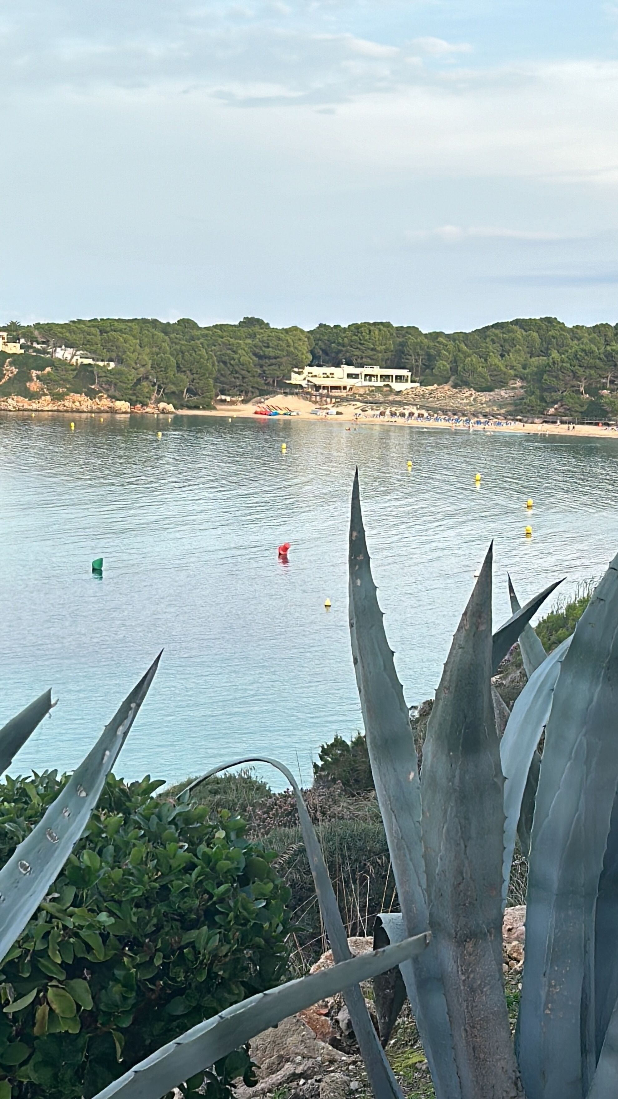 Vista costera con playa, un edificio en una colina y una masa de agua con boyas.