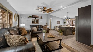 A spacious living room with a leather sofa, a wooden coffee table, and a ceiling fan. A kitchen area is visible in the background with a refrigerator and a dining table. - Secluded cabin close to Eureka Springs and Table Rock Lake (Eureka Springs)
