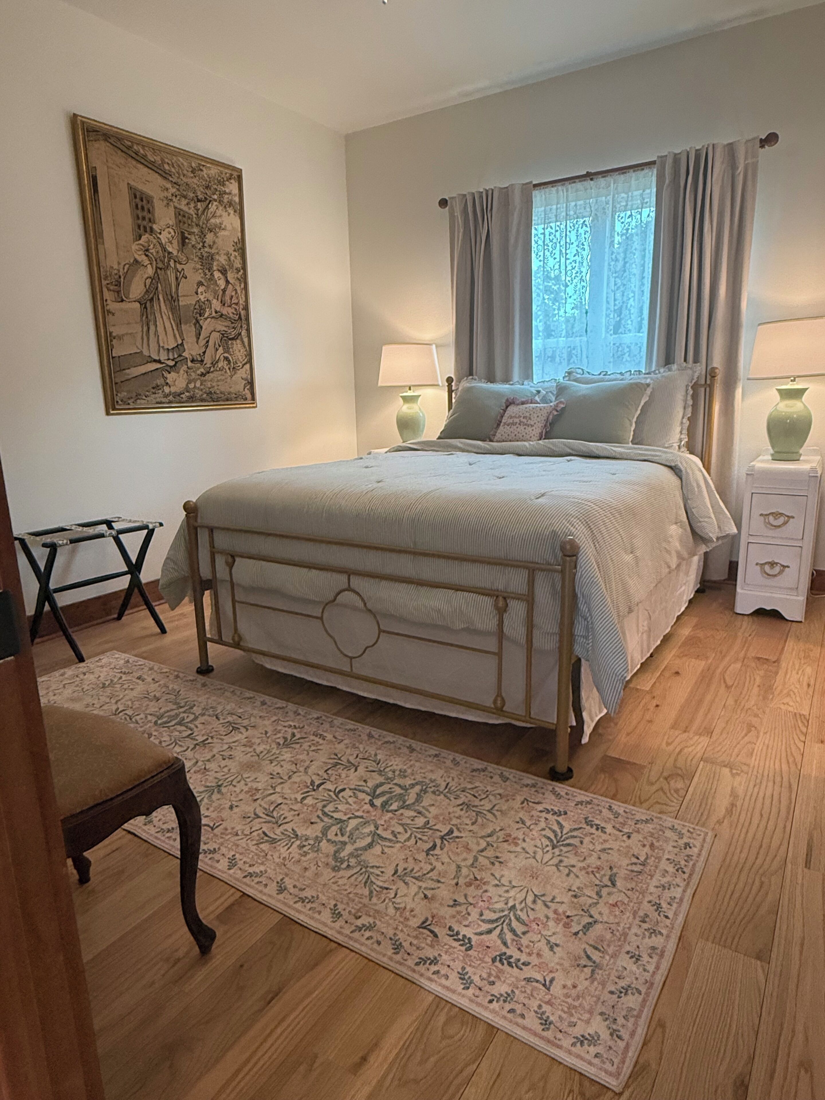 A bedroom with a bed, two bedside tables, a nightstand, a chair, a window with curtains, and a wall-mounted painting.