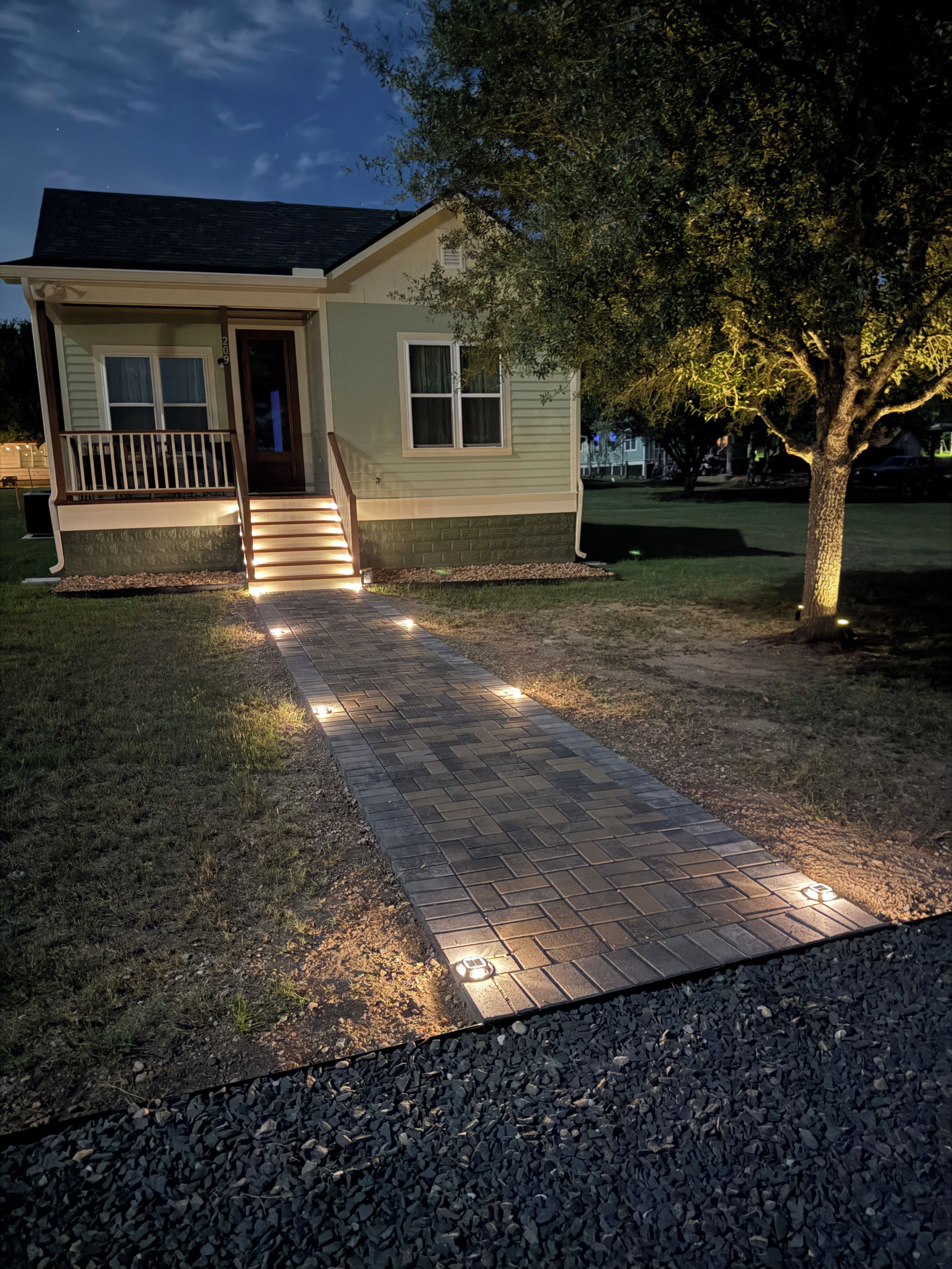 A residential house with a paved driveway leading to the front door, a small porch, and a tree to the right.