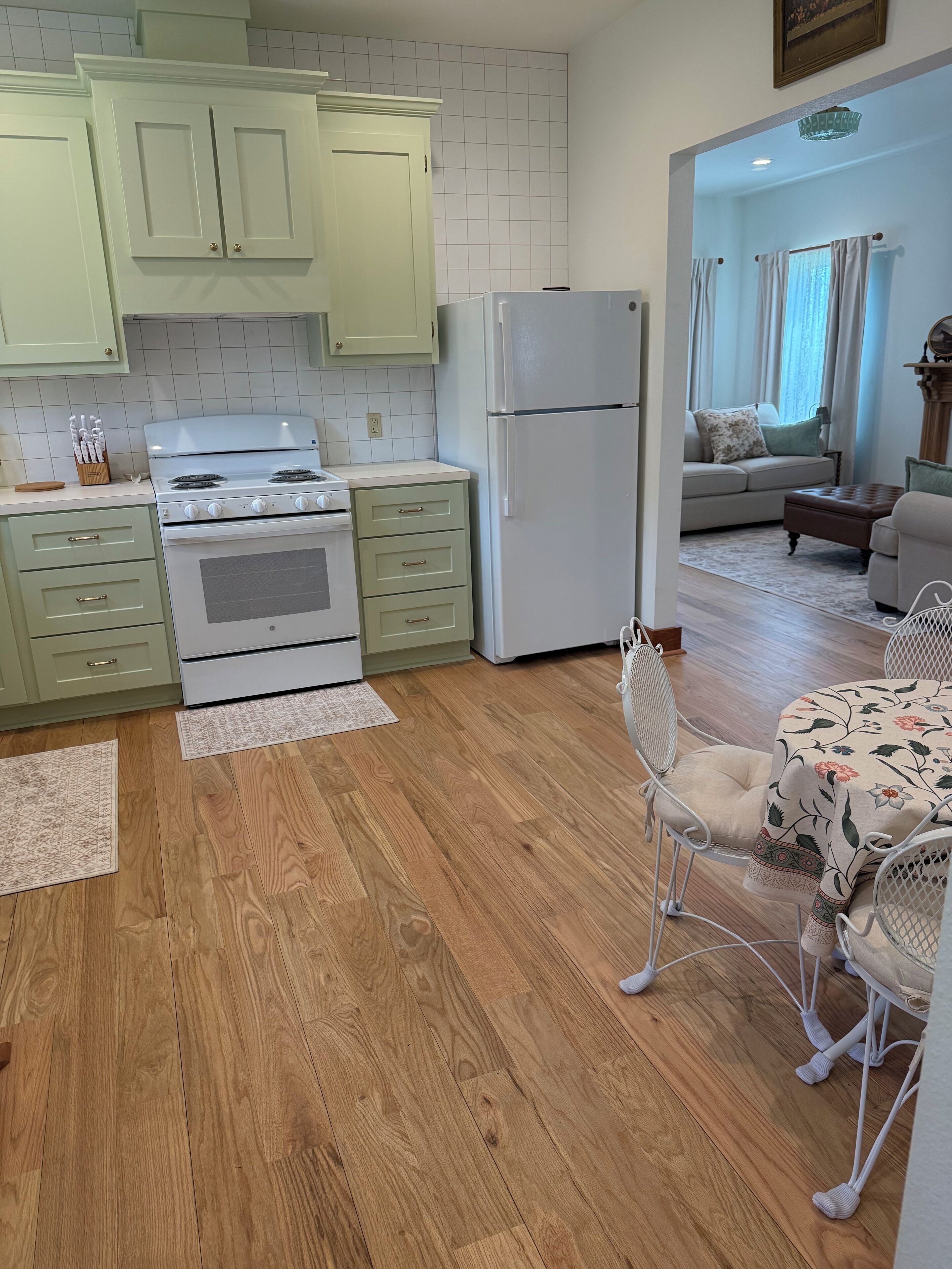 A kitchen with wooden flooring, a dining area with a floral tablecloth, and a living room with a sofa and curtains.