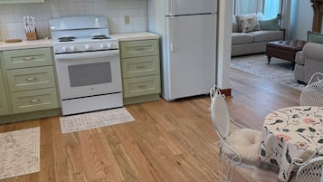 A kitchen with wooden flooring, a dining area with a floral tablecloth, and a living room with a sofa and curtains.