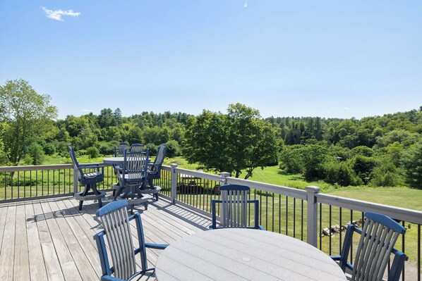Una terraza de madera con mobiliario exterior, barandilla y vista a un paisaje verde.