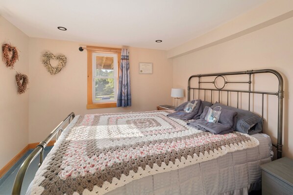 Une chambre à coucher avec un lit en métal, une grande courtepointe, une table de chevet avec une lampe et une fenêtre avec des rideaux.