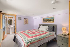 Un dormitorio con cama, mesita de noche, lámpara, un cuadro enmarco y espejo.