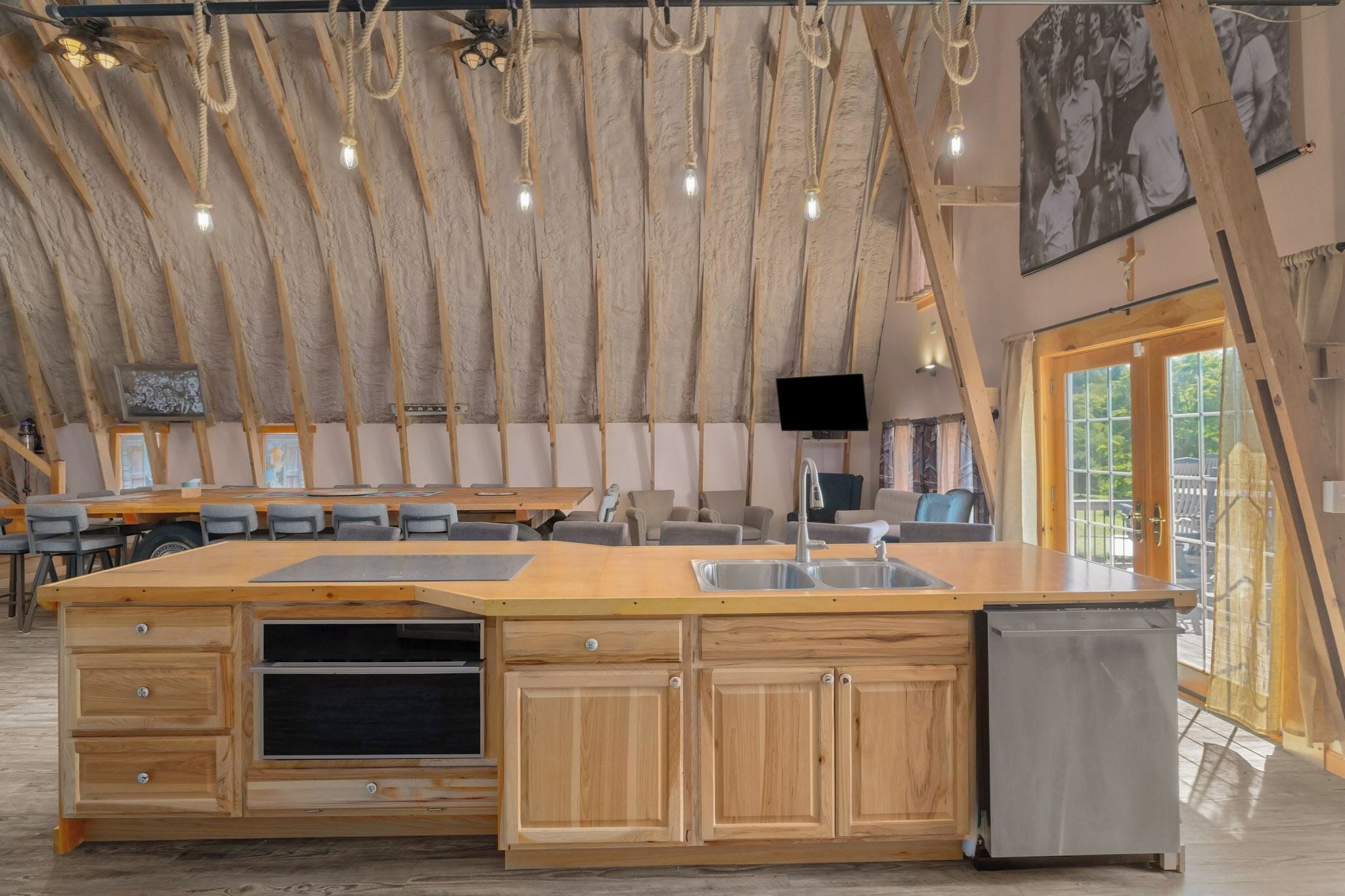 Een ruime keuken met houten kasten, een groot eiland met een spoelbak en een inbouwoven. Er hangt een flatscreen televisie aan de muur en er is een plafondventilator met zichtbare balken.