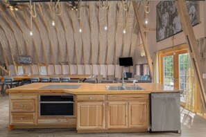 A spacious kitchen with wooden cabinets, a large island with a sink, and a built-in oven. There is a flat-screen TV mounted on the wall and a ceiling fan with exposed beams.