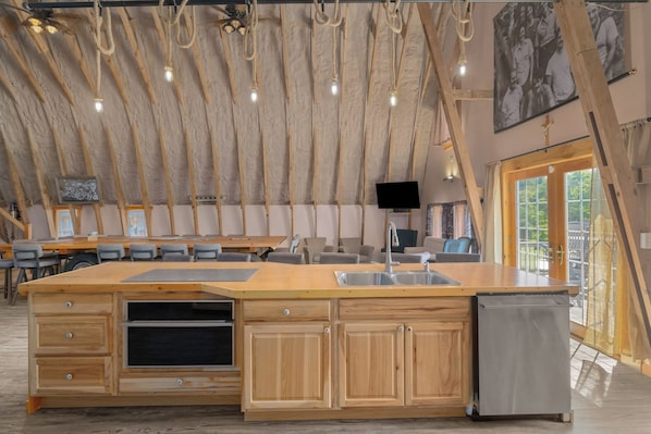 Cocina amplia con armarios de madera, una isla grande con lavamanos y un horno empotrado. Hay un televisor de pantalla plana montado en la pared y un ventilador de techo con vigas a la vista.