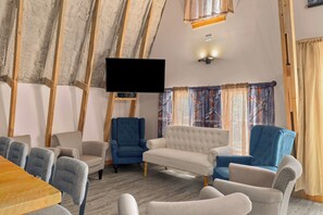 A cozy living room with a flat-screen TV, a wooden ceiling, and a large window with curtains.