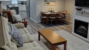 Una sala moderna con sofá, mesa de centro y chimenea. Se observa un área de comedor con mesa y sillas. Se distingue una cocina al fondo.