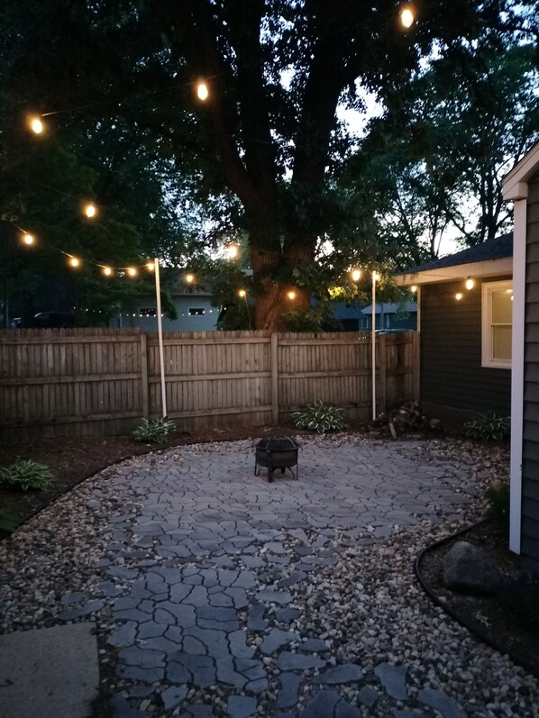 Een achtertuin met een terras van natuursteen, een vuurkorf, lichtslingers en een houten omheining.