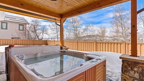 Un bain à remous situé dans un espace de terrasse en bois, avec un mur en pierre et une vue sur une maison et des arbres en arrière-plan.
